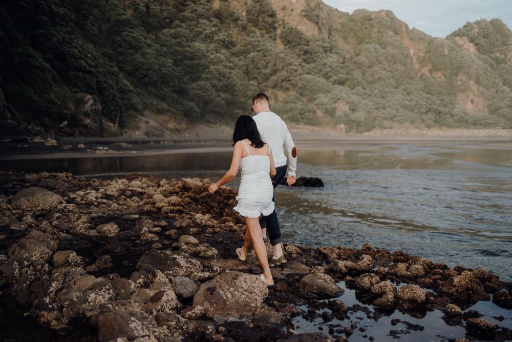 A couple explores a rocky coastline at sunset with mountains in the background.