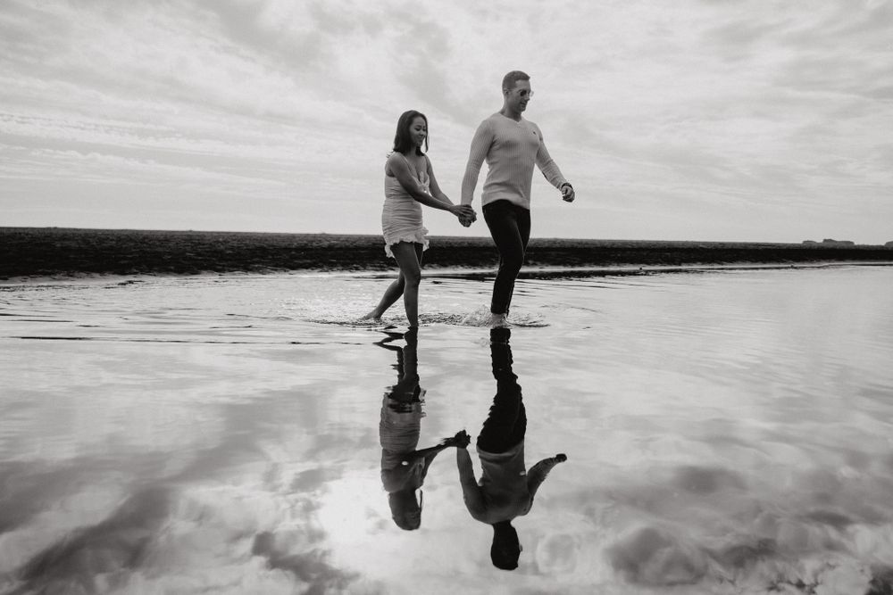 A couple walks playfully in shallow water creating mirror reflections at the beach.
