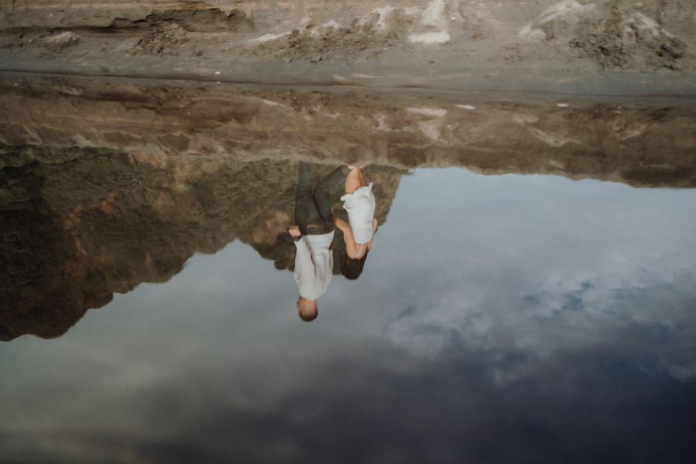 A couple's reflection appears in still water against a dramatic rocky landscape.