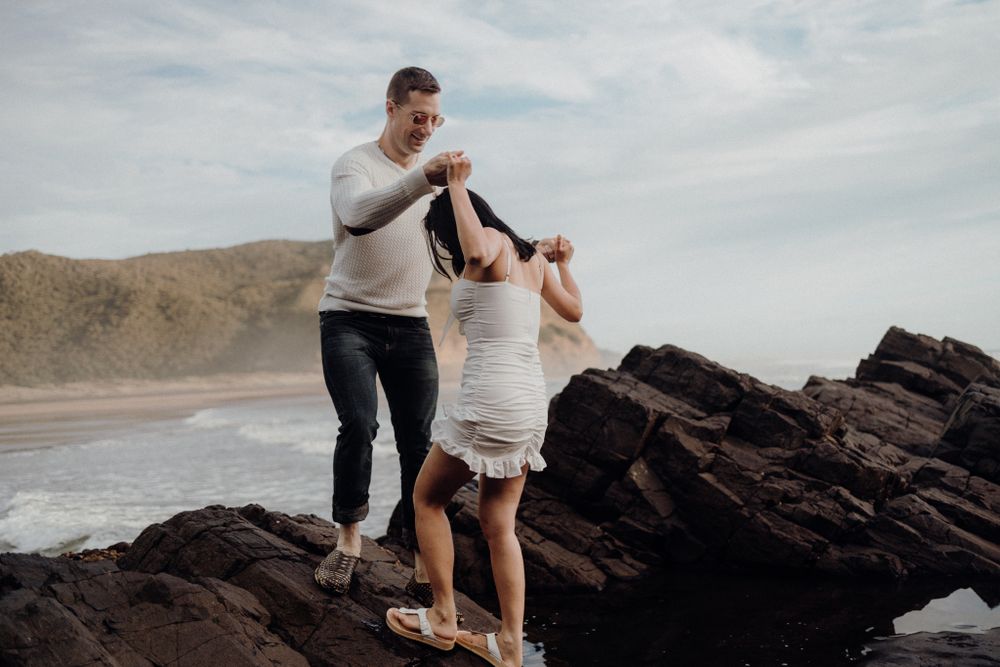 A couple navigates rocky terrain together along a scenic coastal backdrop.