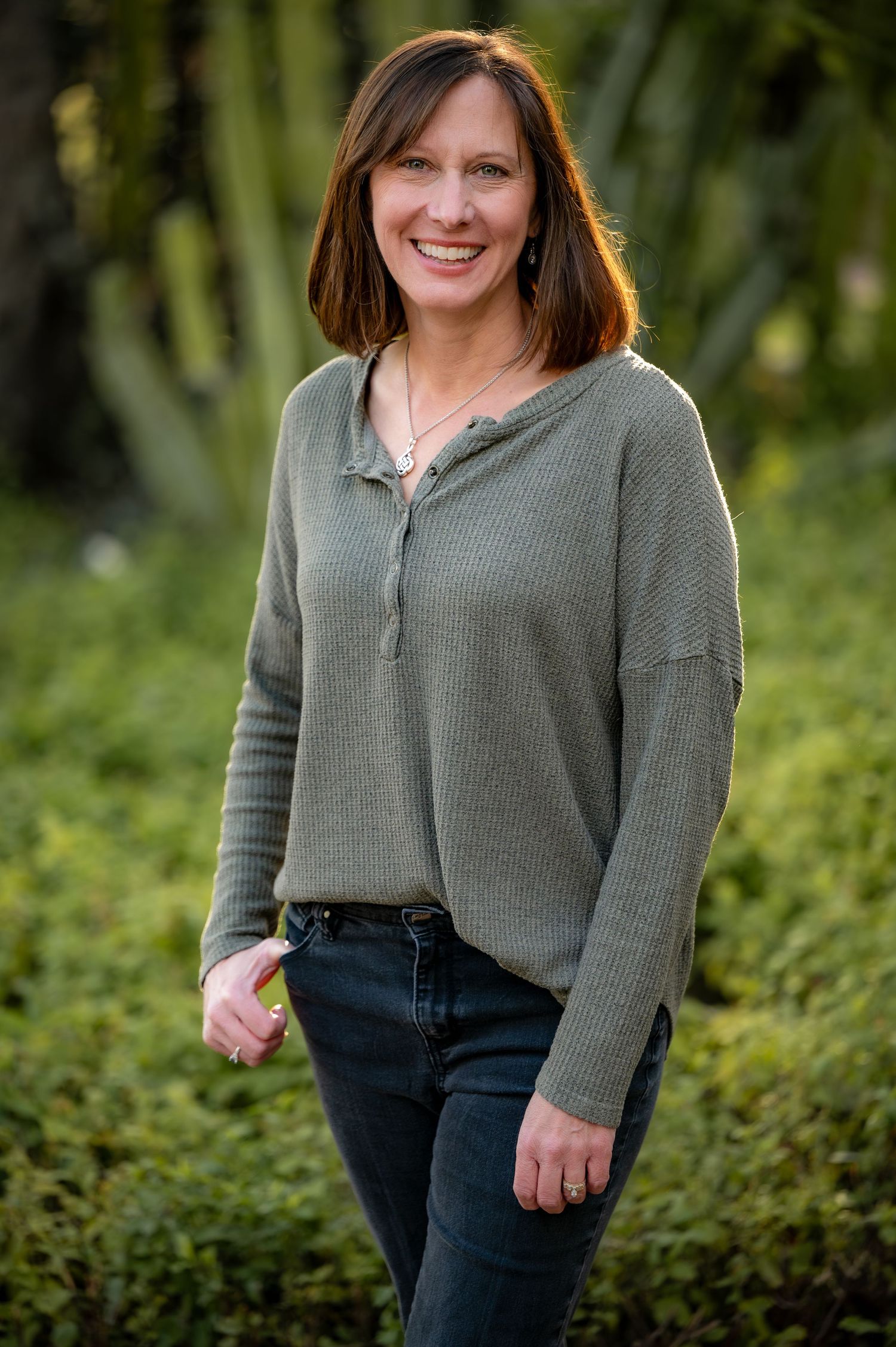Woman in gray sweater and jeans smiles warmly outdoors against natural foliage background.
