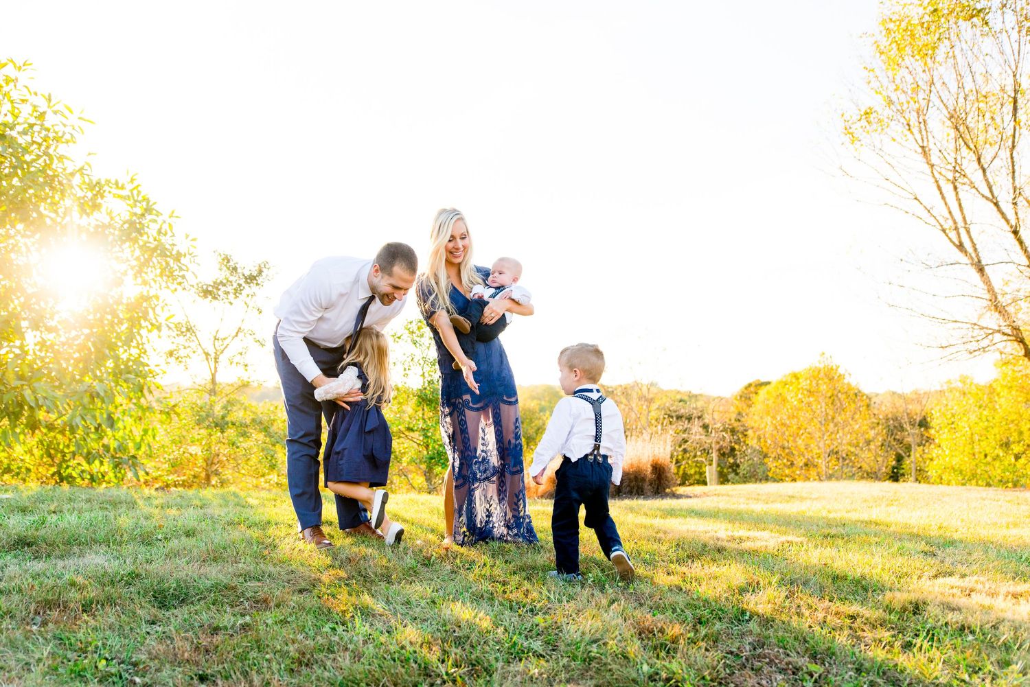 mother holding baby and daughter hugging father as other little boy walks toward family in navy and white clothes