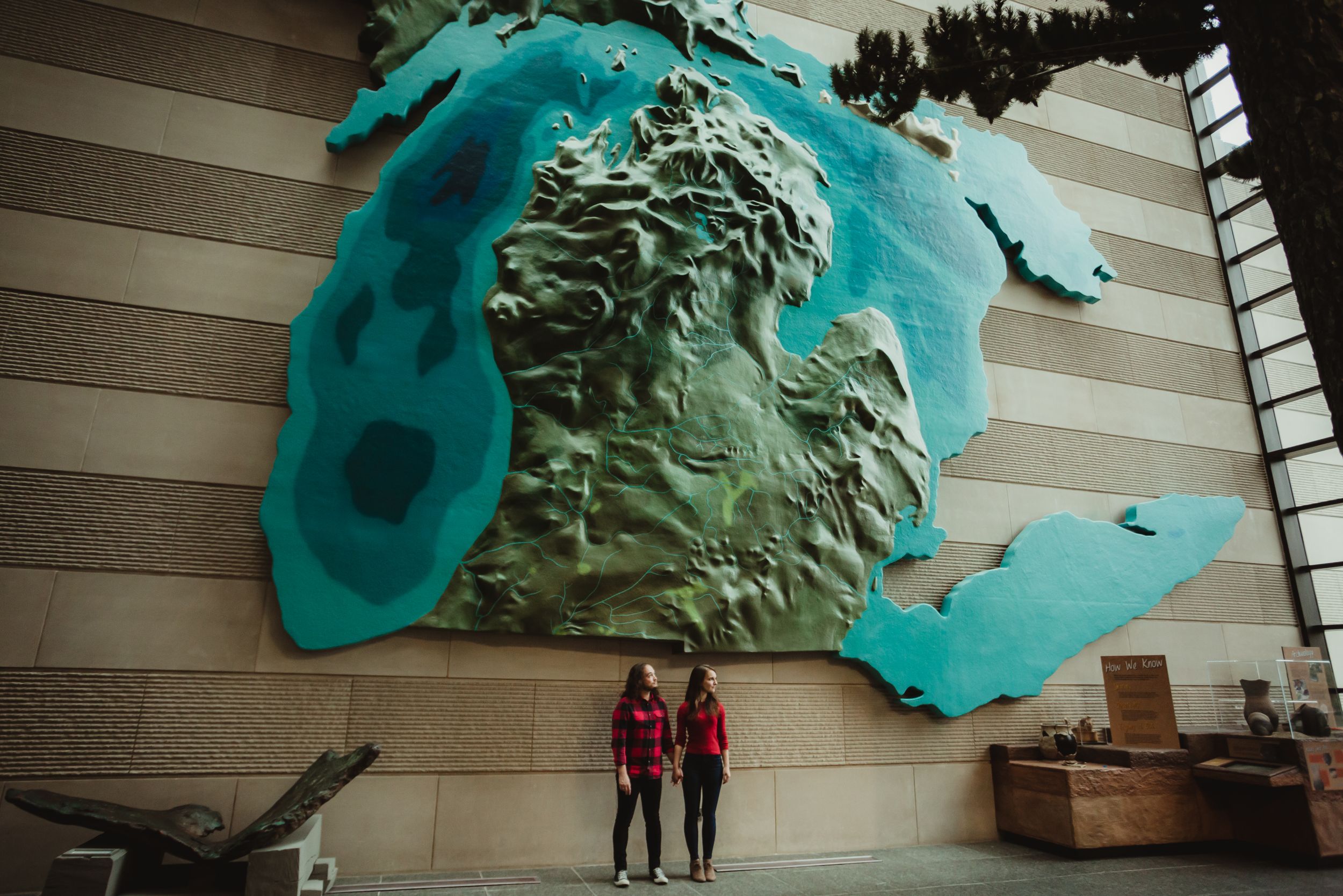 Engaged man and woman with brown hair and red shirts standing below are large sculpture of Michigan.