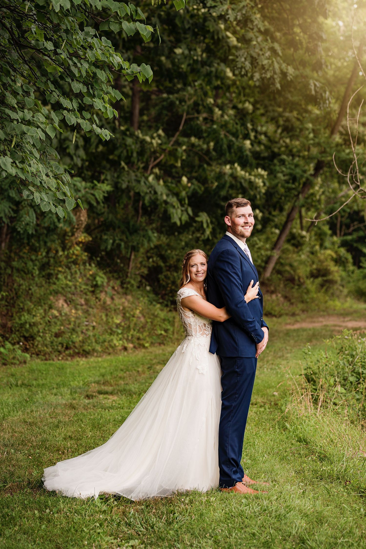 Joyful wedding couple poses back-to-back on grassy path with sunlit forest background.
