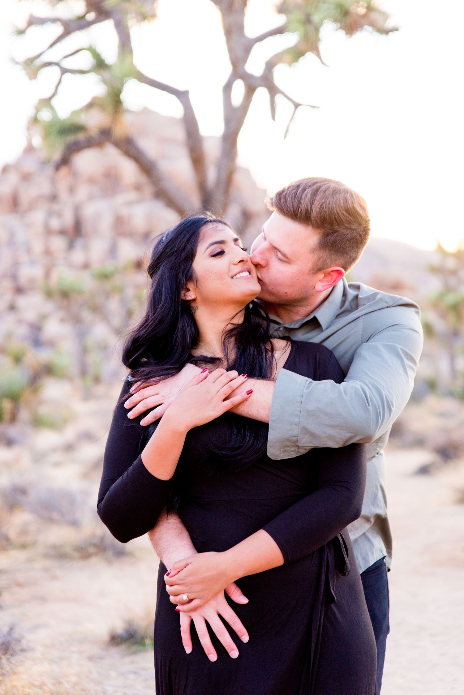 man in gray shirt kissing his fiancee in a black dress while hugging her from behind at Joshua Tree National Park