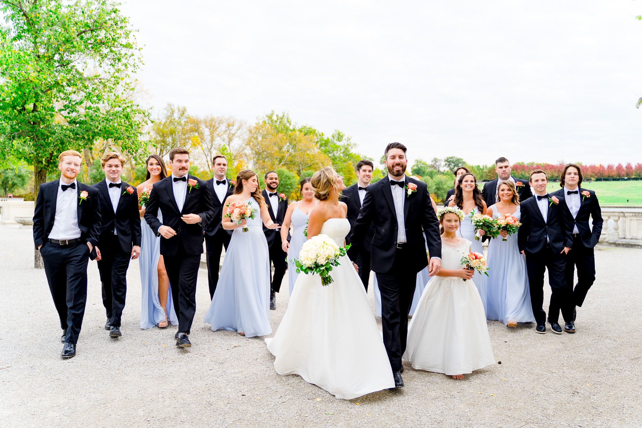 large wedding party in black suits and light blue dresses walk with bride and groom at Forest Park STL fall wedding