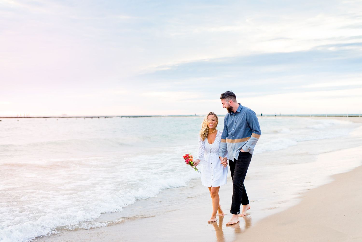 engaged man and woman hold hands and laugh while they walk along the water at North Avenue Beach