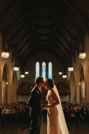 Couple stands at altar in church with stained glass windows and gathered guests in pews.