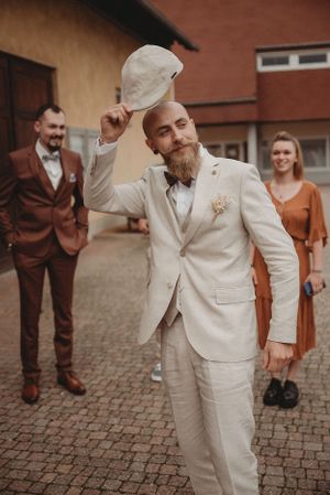 A groom in a light beige suit tips his flat cap while posing outdoors on cobblestones.