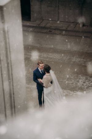 Couple in wedding attire sharing intimate moment in moody concrete setting with falling snow effect.