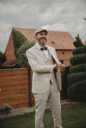 Wedding guest in cream colored suit and bowtie poses in garden setting.