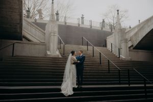 A wedding couple embraces on grand stone steps during an overcast day.