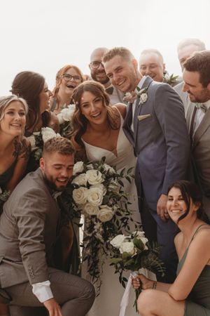 Wedding party gathers for joyful group photo with white floral bouquet.