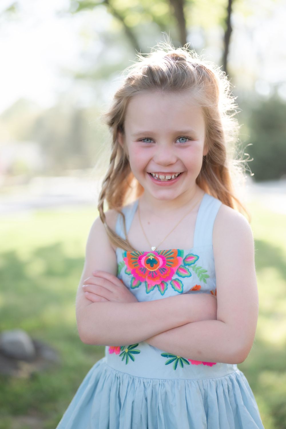 Young child in light blue dress with floral design smiles brightly against blurred outdoor background.