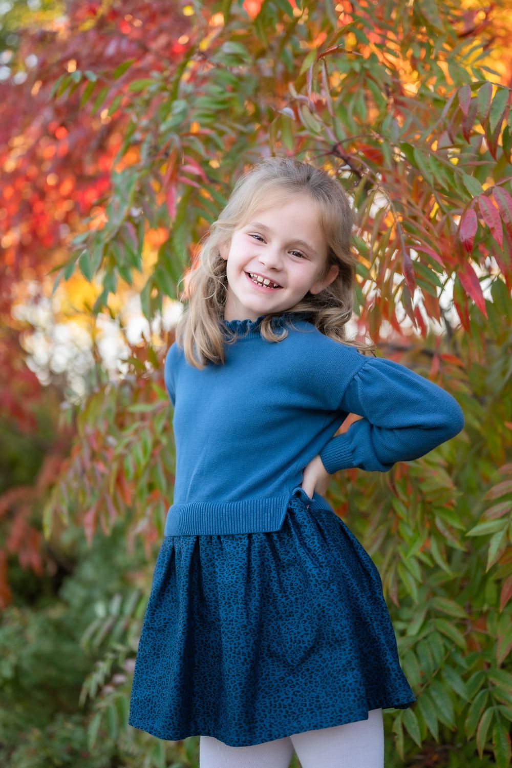 Child poses playfully in blue outfit against vibrant autumn foliage background.