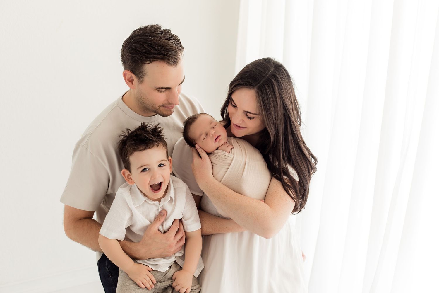Mom and dad staring at their toddle and newborn baby next to a bright white window room for their family newborn portrait session.