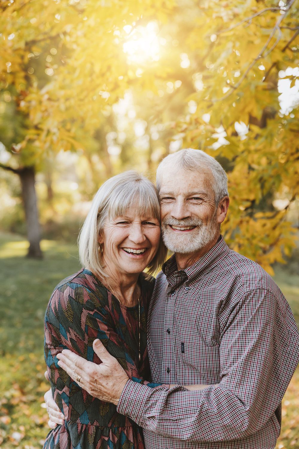 An older couple poses in the sun with yellow autumn leaves.