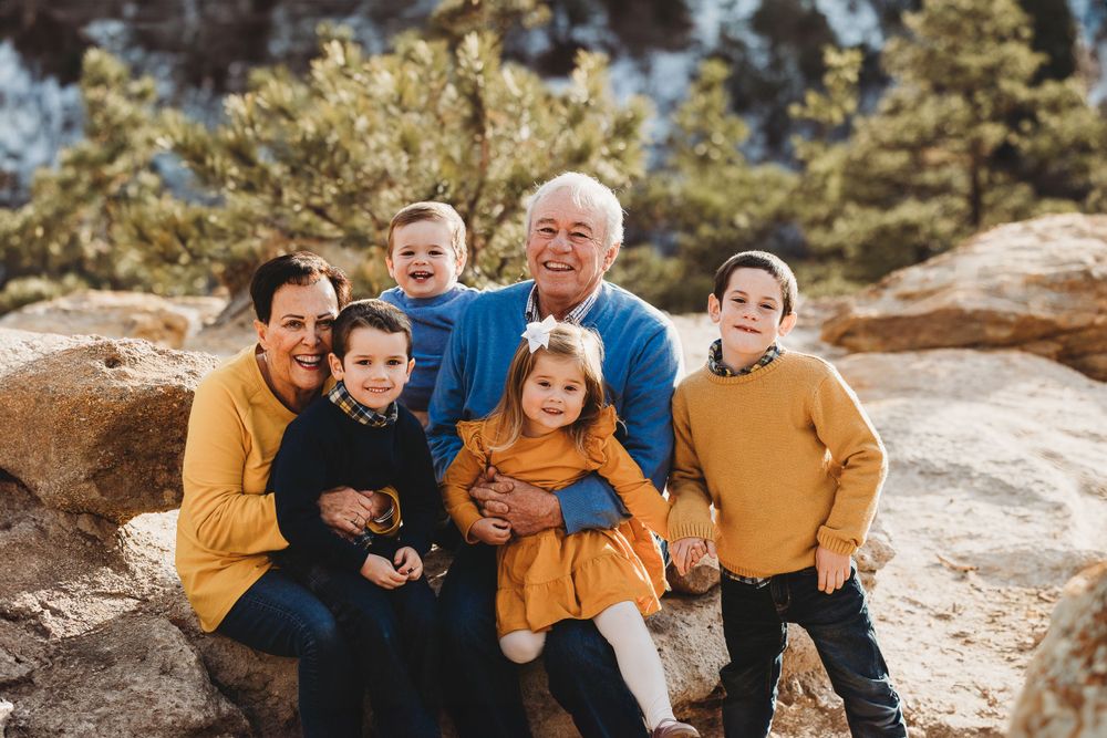 Grandparents and their grandchildren wear yellow and blue sweaters and sit on rocks in the sun for a portrait.