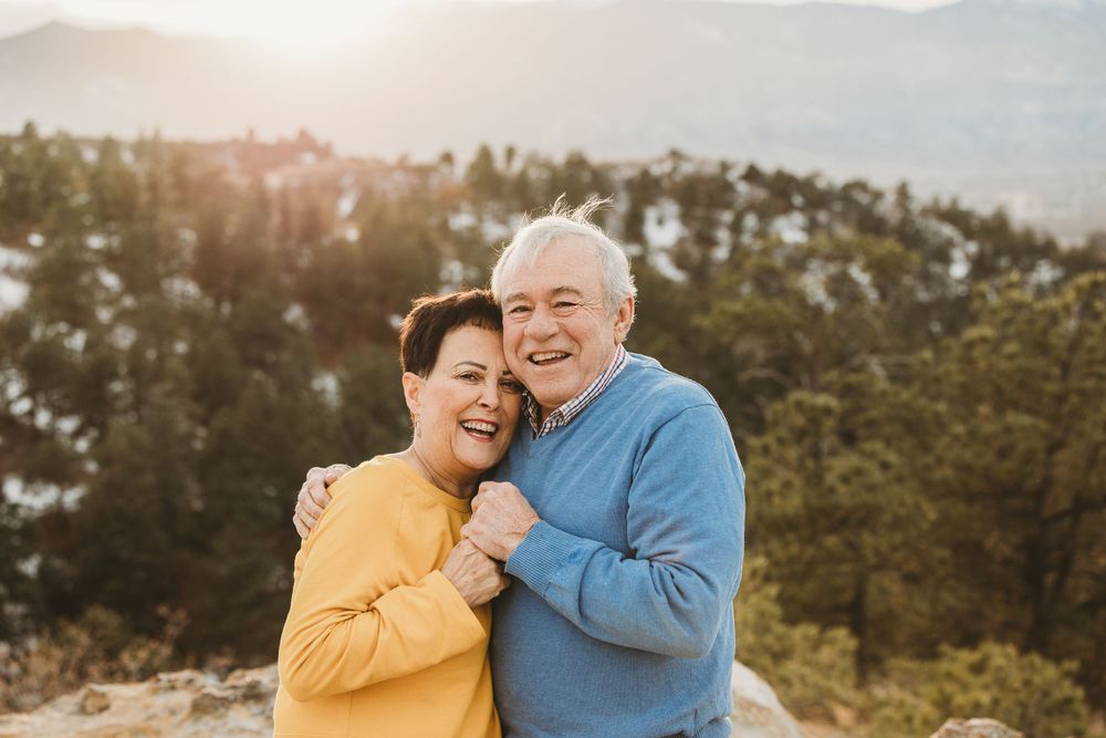 Two elderly people embrace in the sunlight with a snow forest at golden hour.