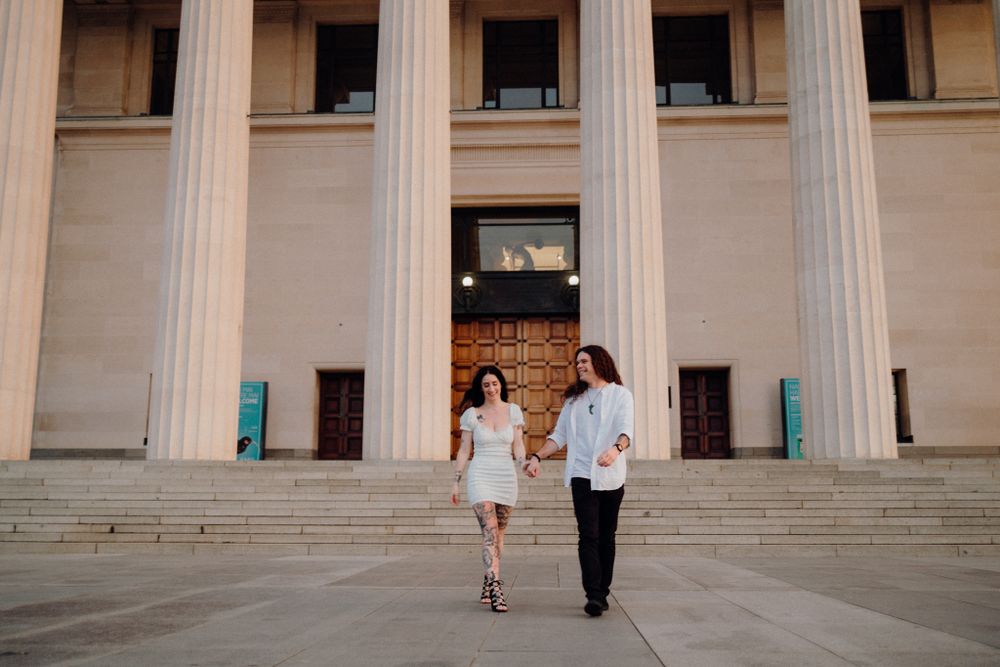A couple descends stone steps while holding hands in front of a columned building entrance.