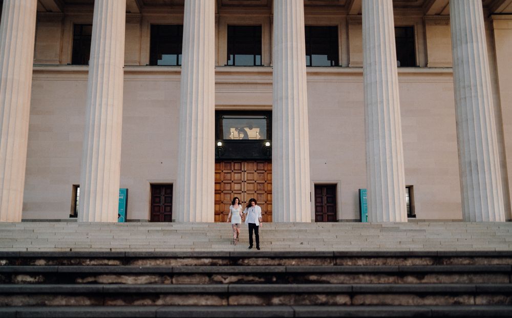 A couple stands on stone steps between grand white columns of a neoclassical building facade.