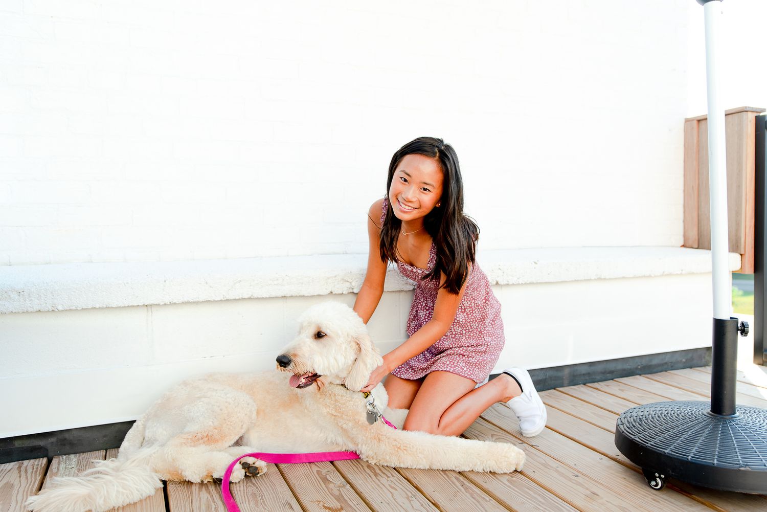 high school senior girl kneeling down to pet her dog in front of a white wall