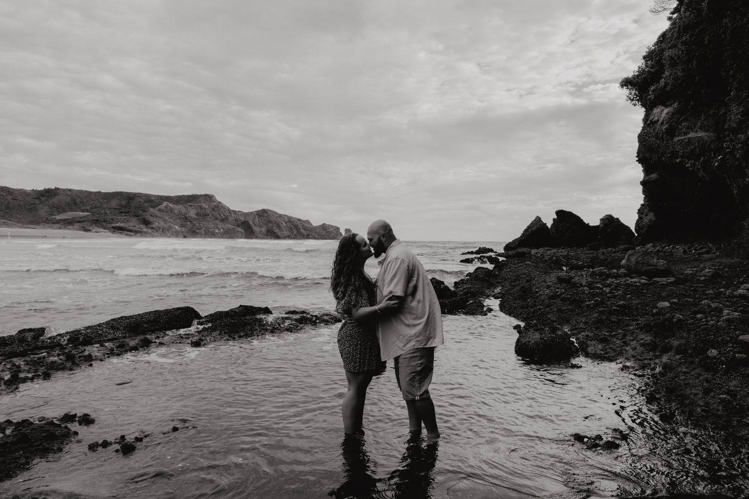 engaged couple stand in a rock pool up to their ankles and kiss