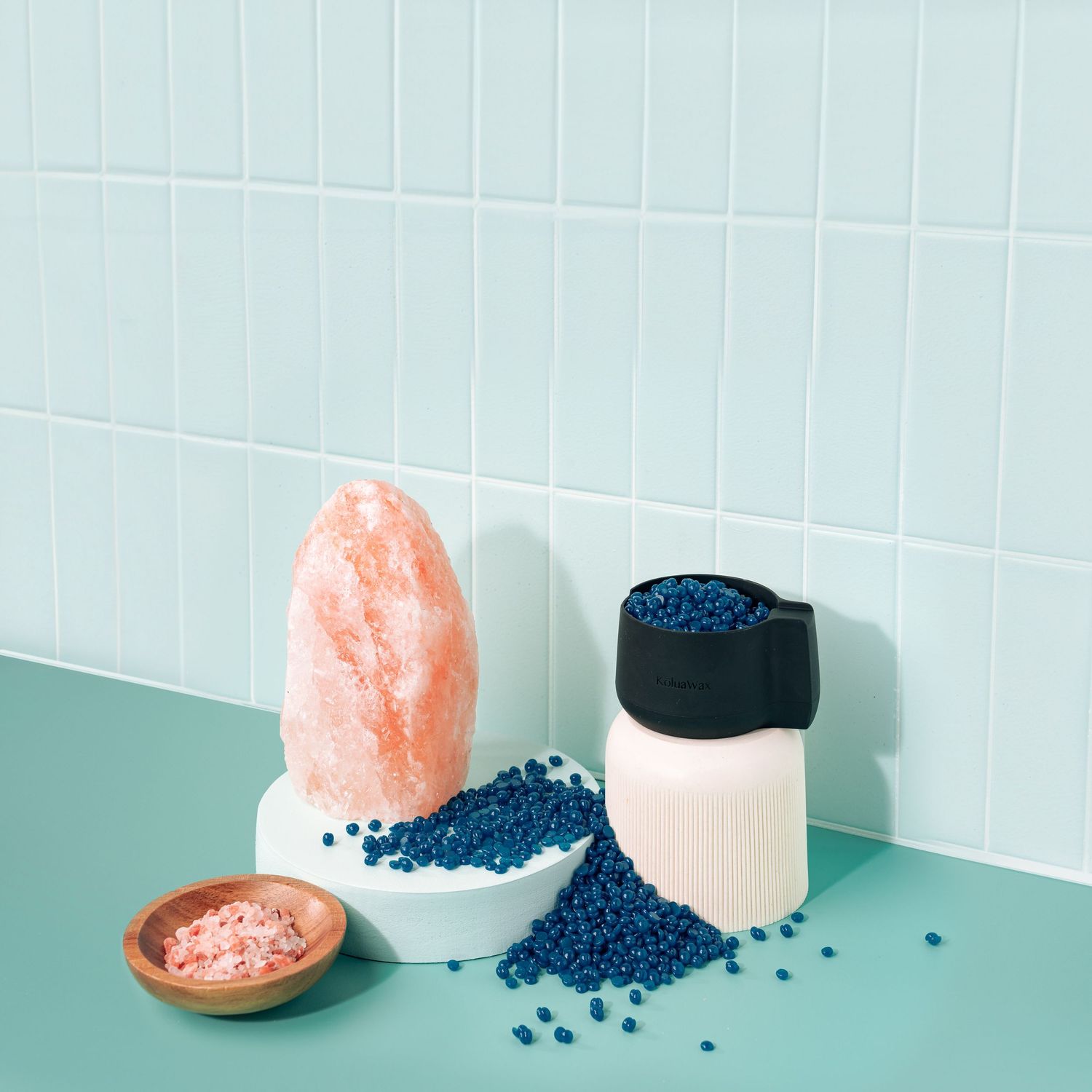 Pink salt crystals and blue beads displayed with ceramic containers against white tile wall.