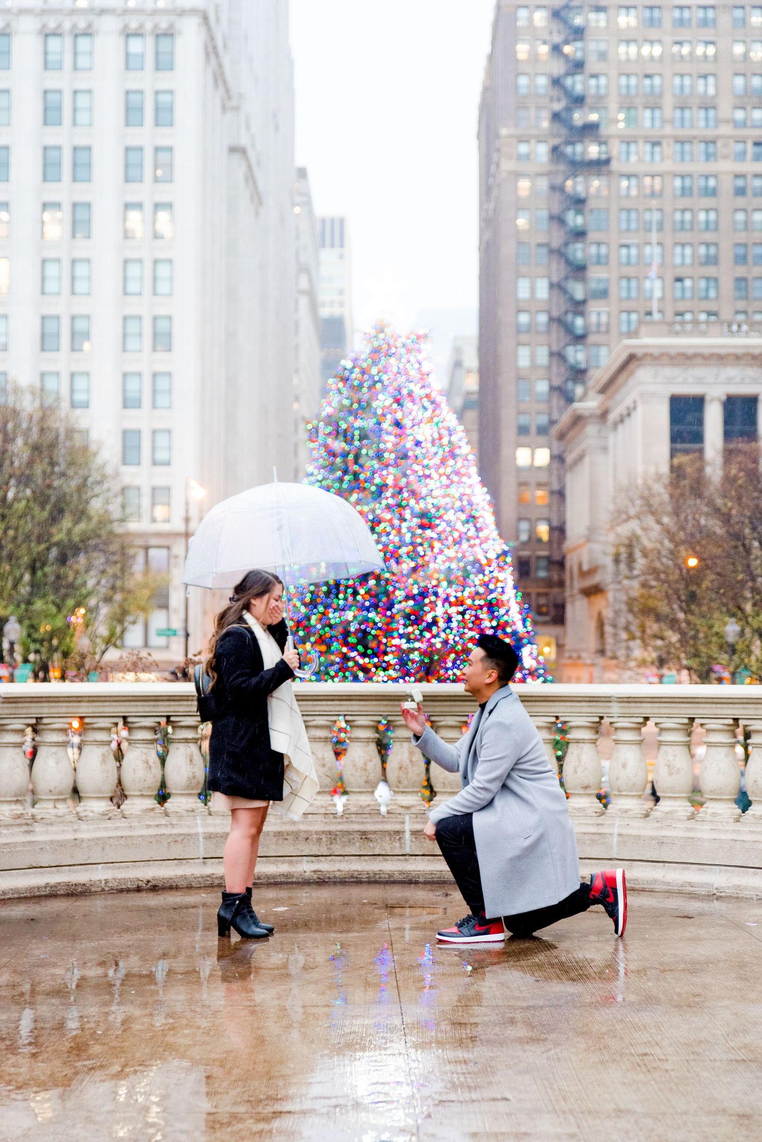 man in grey coat kneeling proposing to woman in black coat and clear umbrella in front of Millennium Park Christmas Tree
