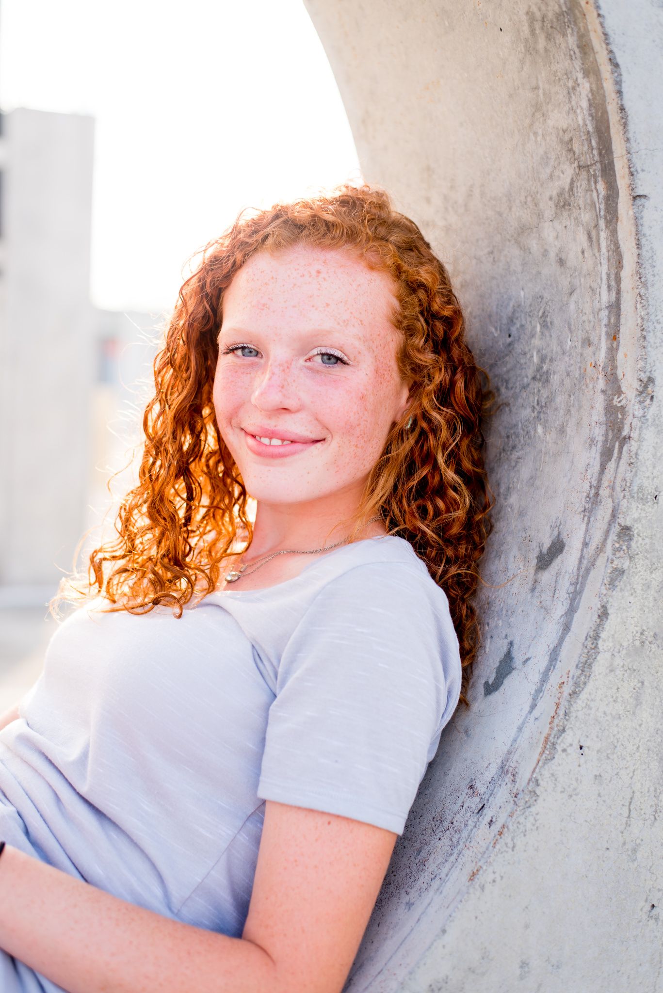 young woman with red curly hair leaning against concrete circle cut out, smiling at sunset in downtown CIncinnati
