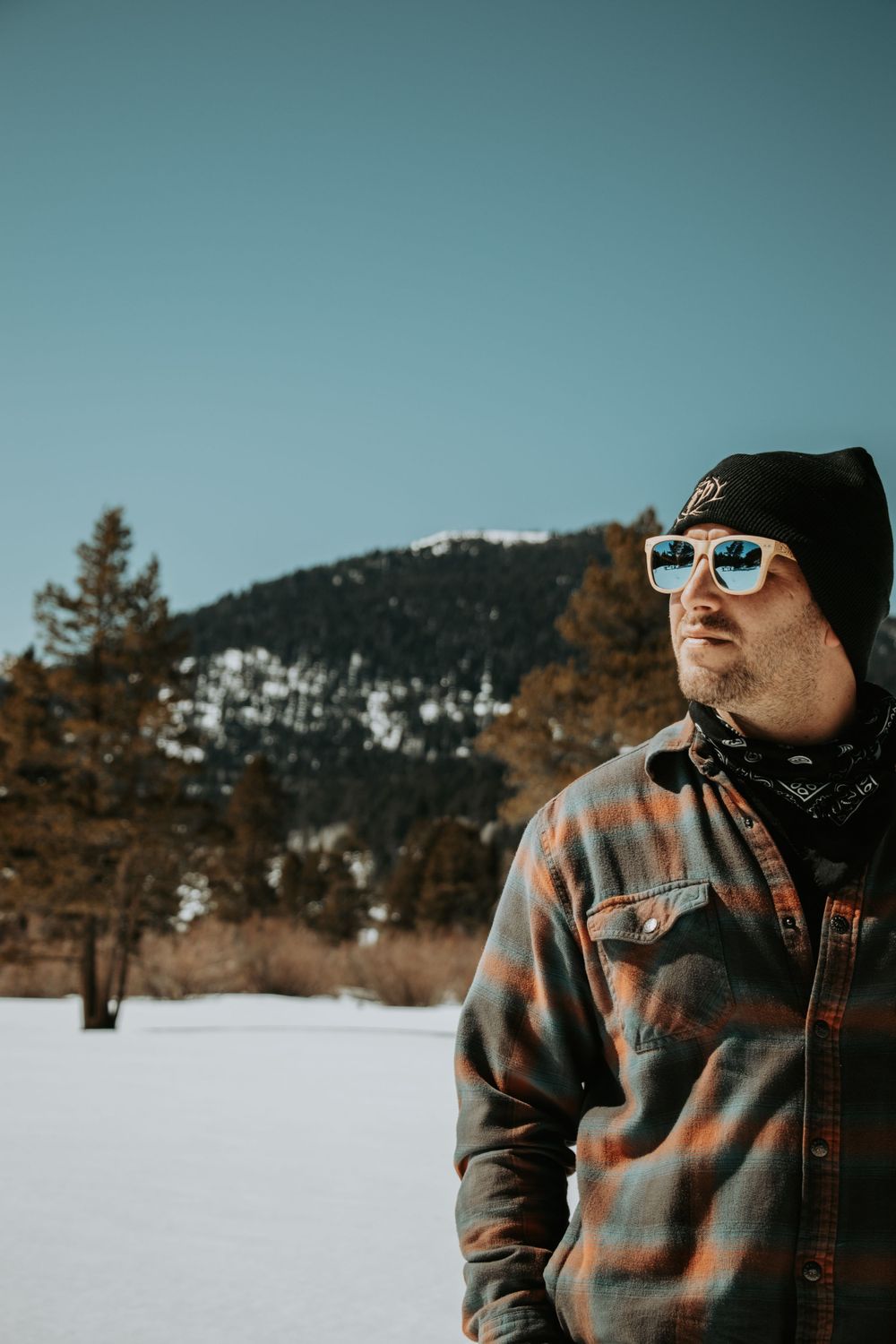 Person wearing sunglasses and winter clothing standing in snowy landscape with mountain backdrop.
