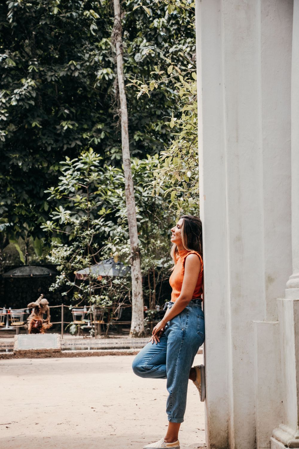 Woman in orange top and jeans leaning against white pillar in sunny outdoor setting.