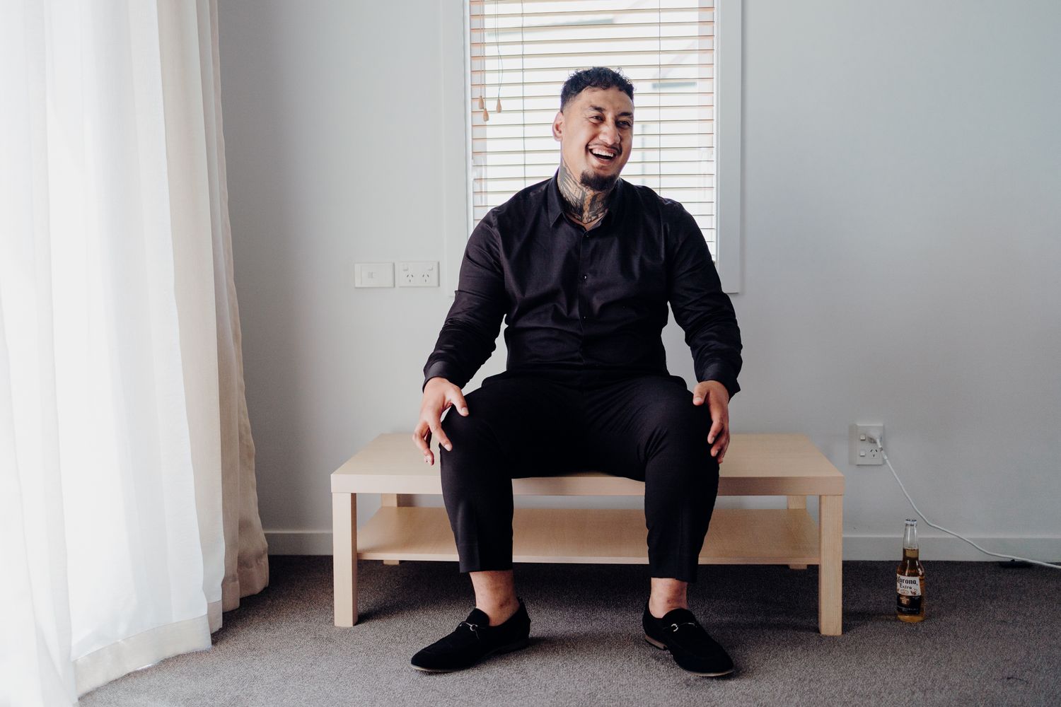 Person in black outfit sitting casually on wooden bench near window.