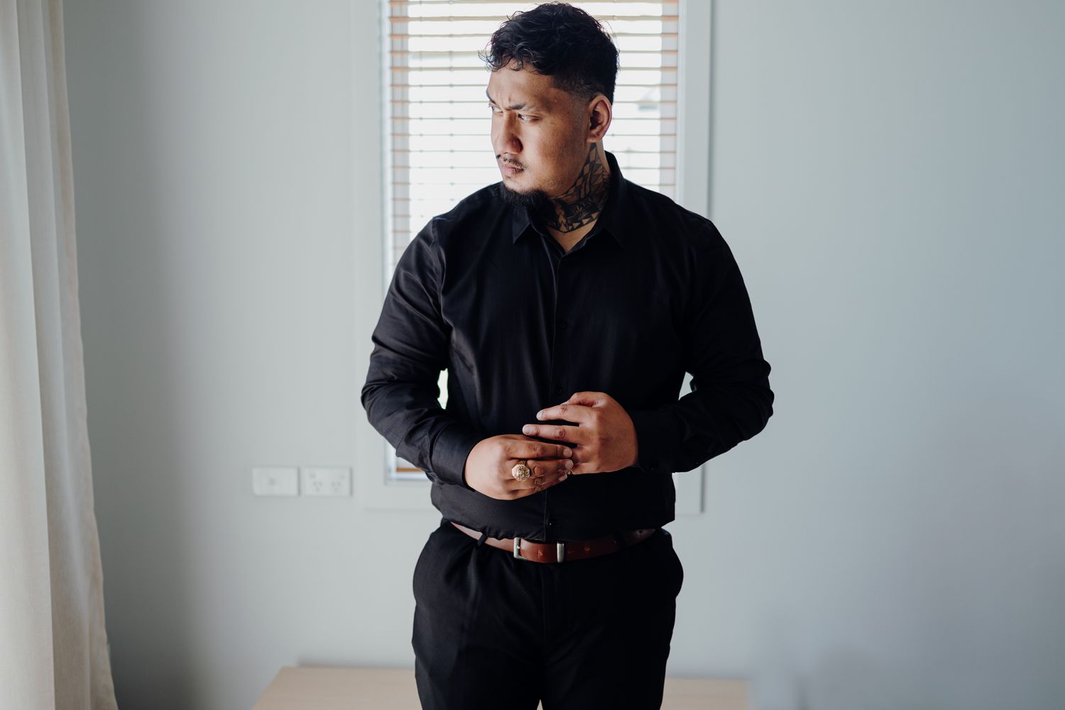 A person in formal black attire adjusts their sleeves while standing near a window with venetian blinds.
