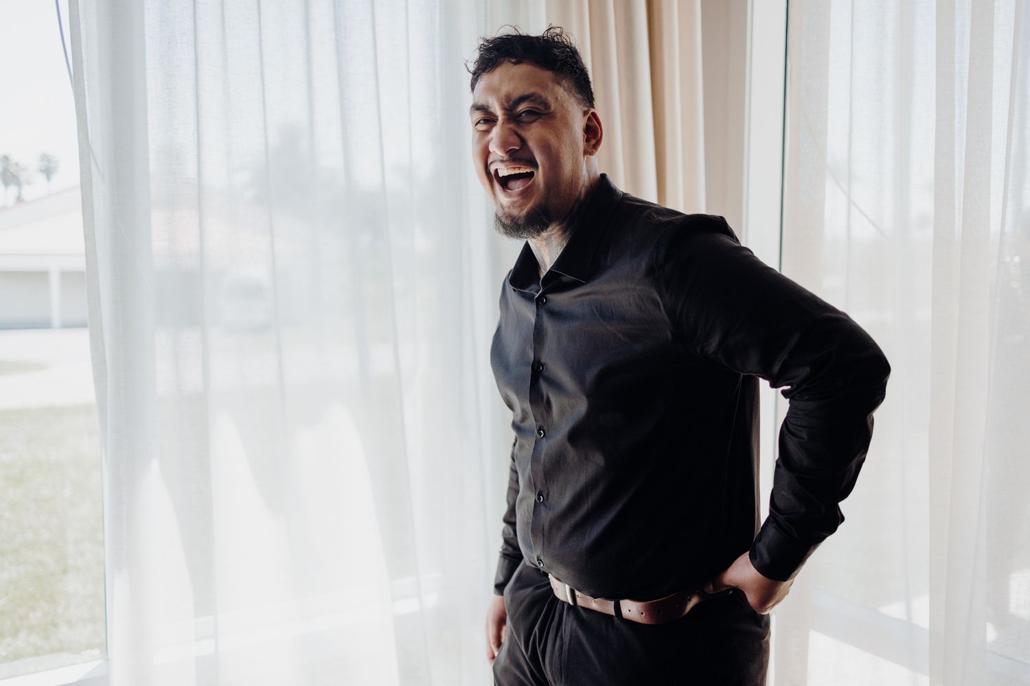 Person in formal black attire smiling in front of sheer curtains.
