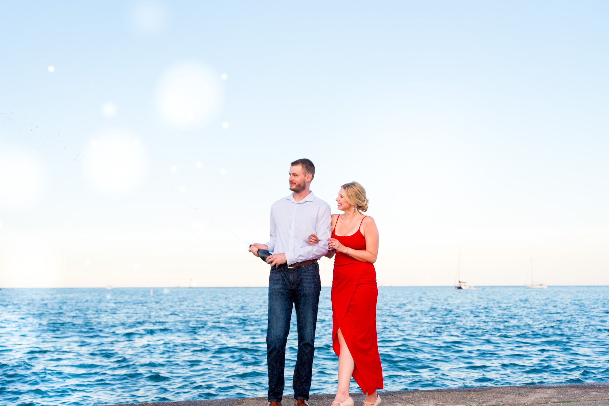 man popping bottle of champgne while woman in red dress smiles next to him watching