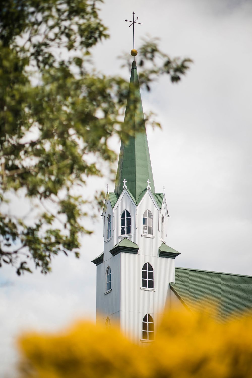 Tall white church steeple with green roof rising above yellow flowers against cloudy sky.