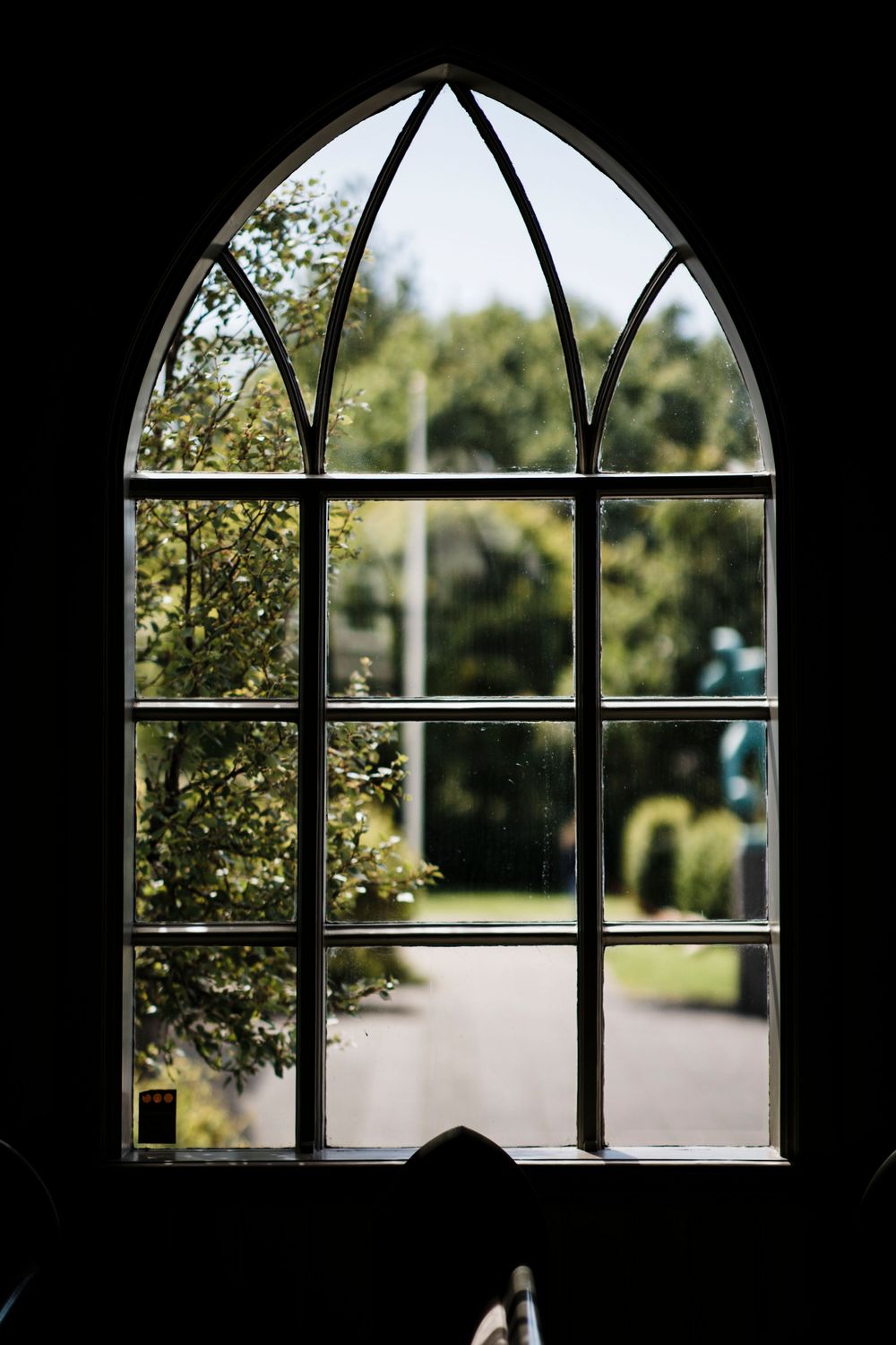 Gothic arched church window frames a serene garden view with sunlit trees.
