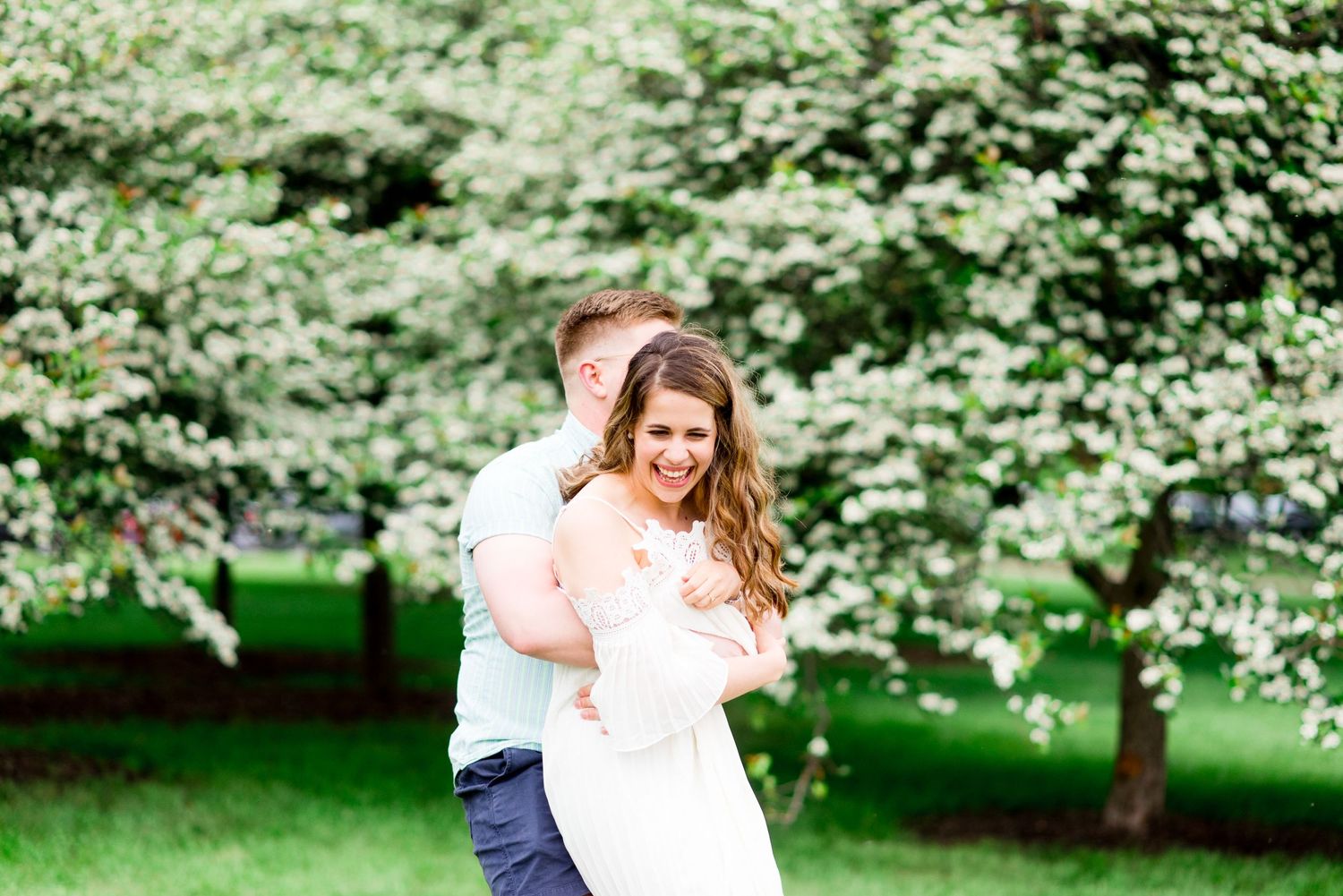 engaged couple playfully hugs and spins around in front of white flower bush at Chicago Montrose Harbor