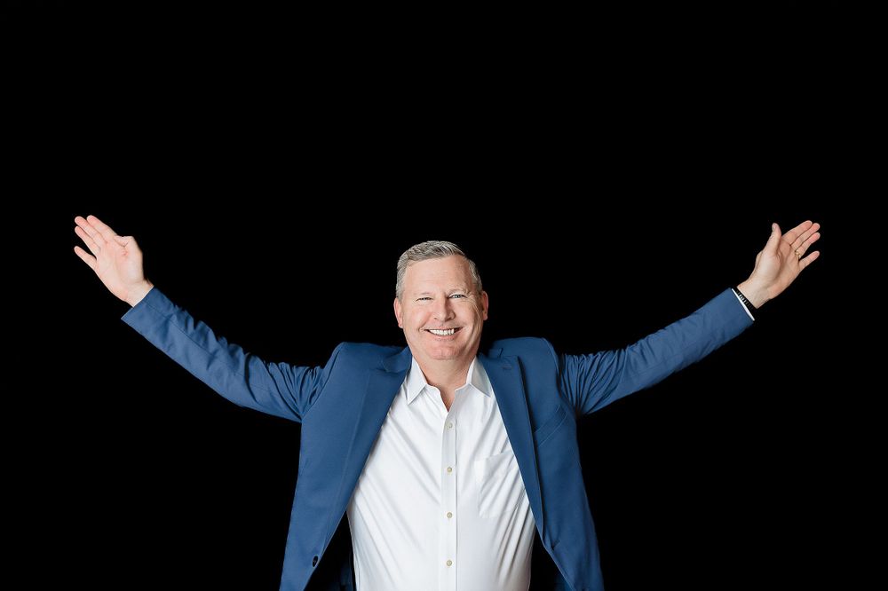Man in blue blazer spreads arms wide against black background with joyful expression.
