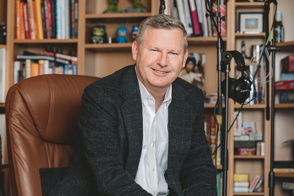 Professional studio portrait of a person in dark blazer and white shirt seated in a leather chair with bookshelves behind.