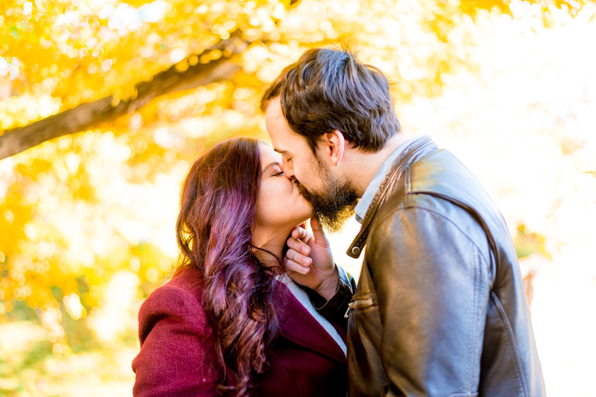 brown hair man kissing red hair woman in red coat under yellow trees in Central Park in the fall at sunrise