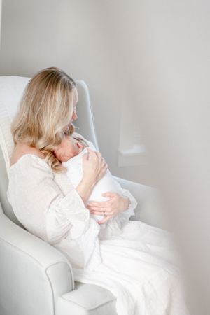 Mother sits in rocking chair holding her newborn baby in all white