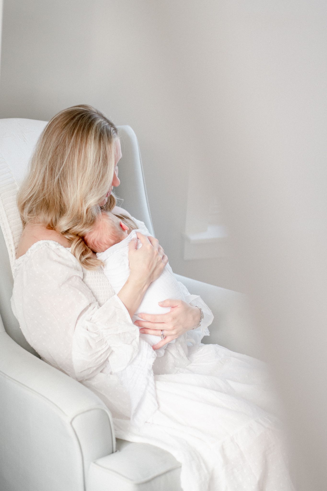 Mother sits in rocking chair holding her newborn baby in all white