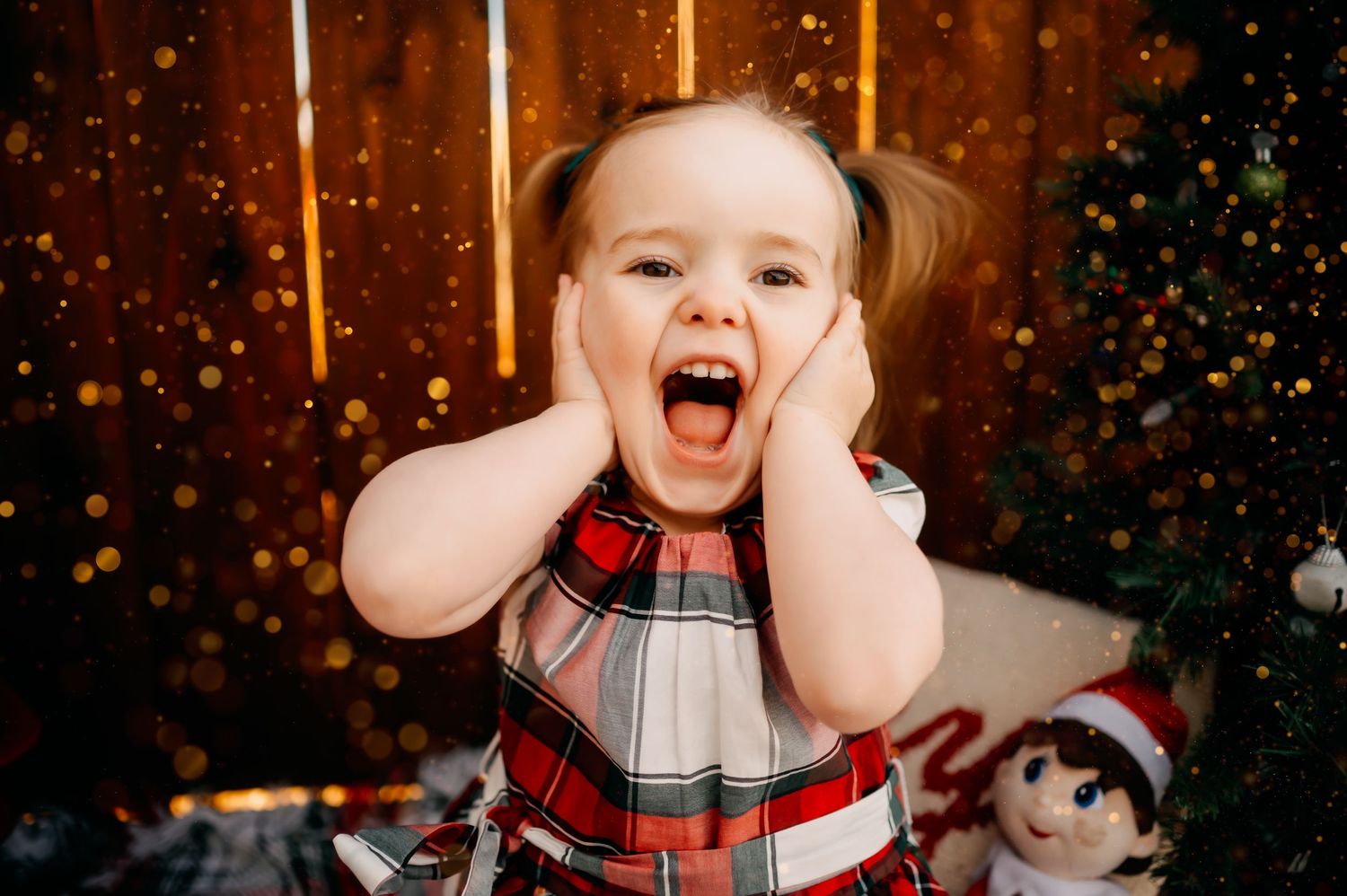 Excited child in plaid holiday dress covering cheeks with hands while sitting near Christmas decorations.