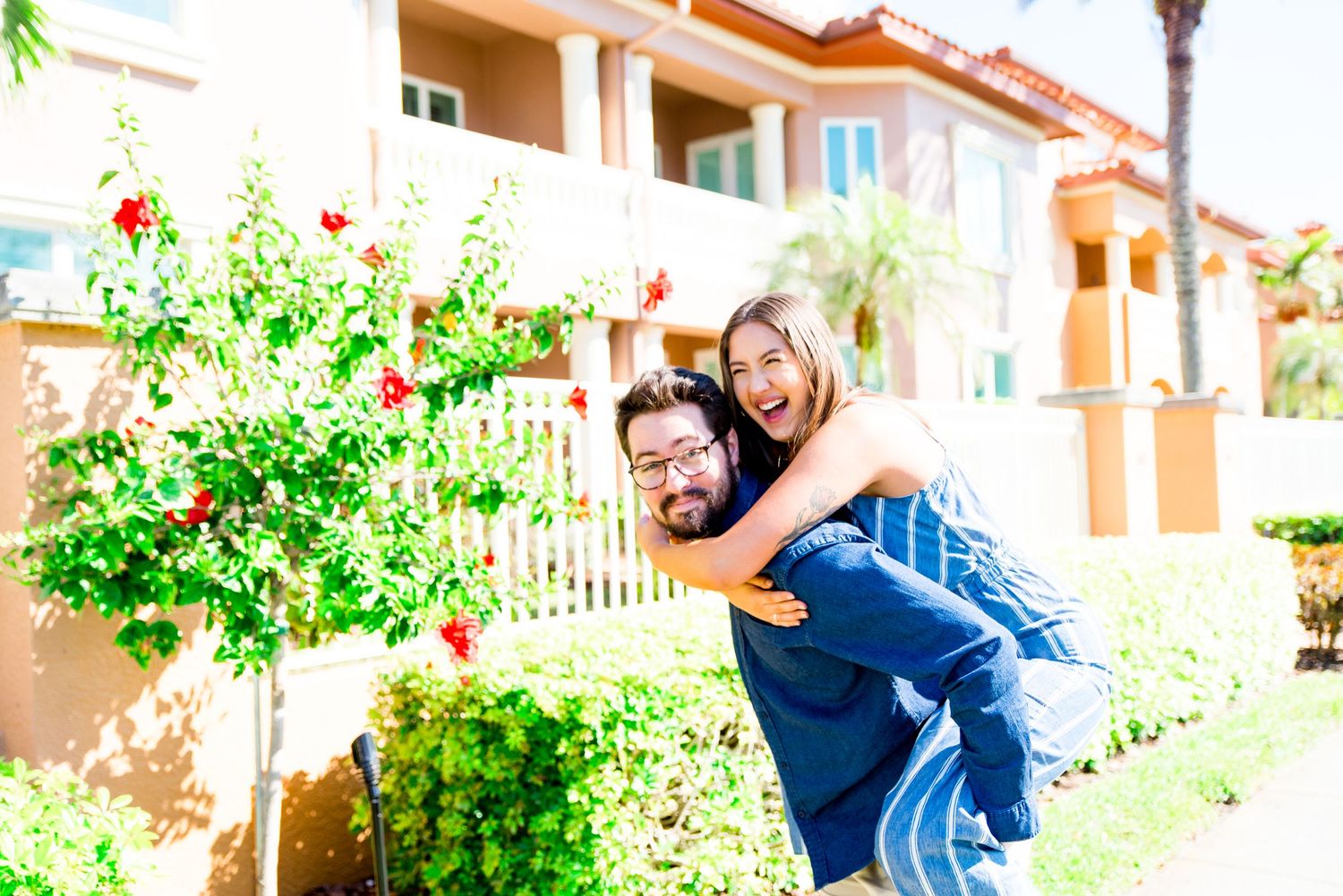 young woman piggyback on boyfriend, laughing next to pink hibiscus tree at Vinoy Park sunrise engagement pictures