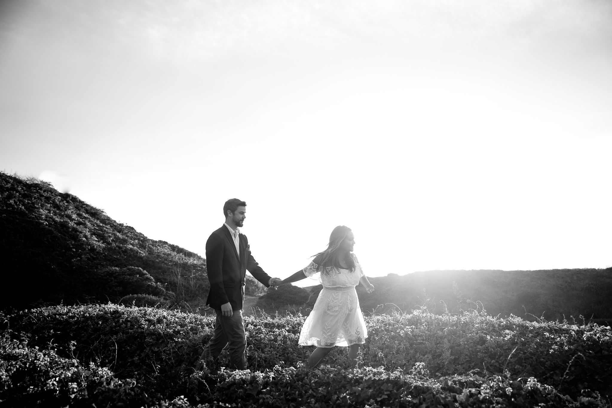 woman in white lace dress leading man in suit through San Francisco hills at sunset engagement pictures