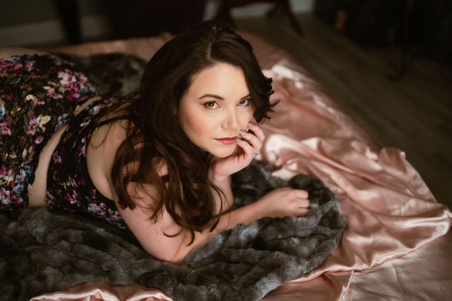 a woman in floral lingerie lies on her front on a bed, gazing into the camera