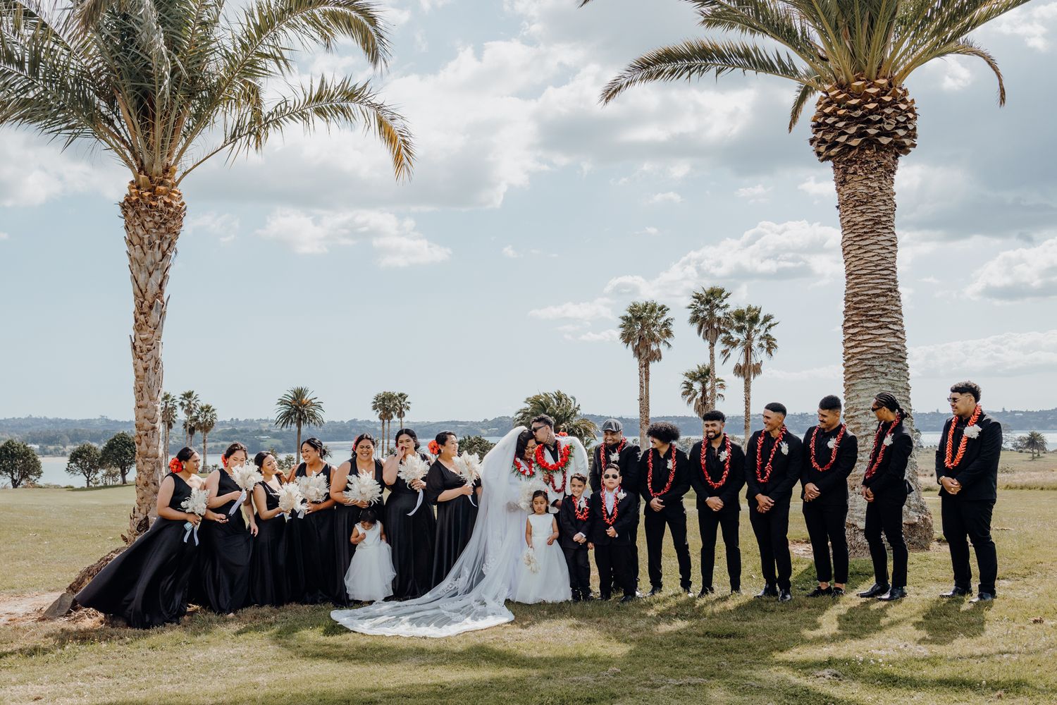 Wedding party standing in a row under palm trees against an ocean backdrop.