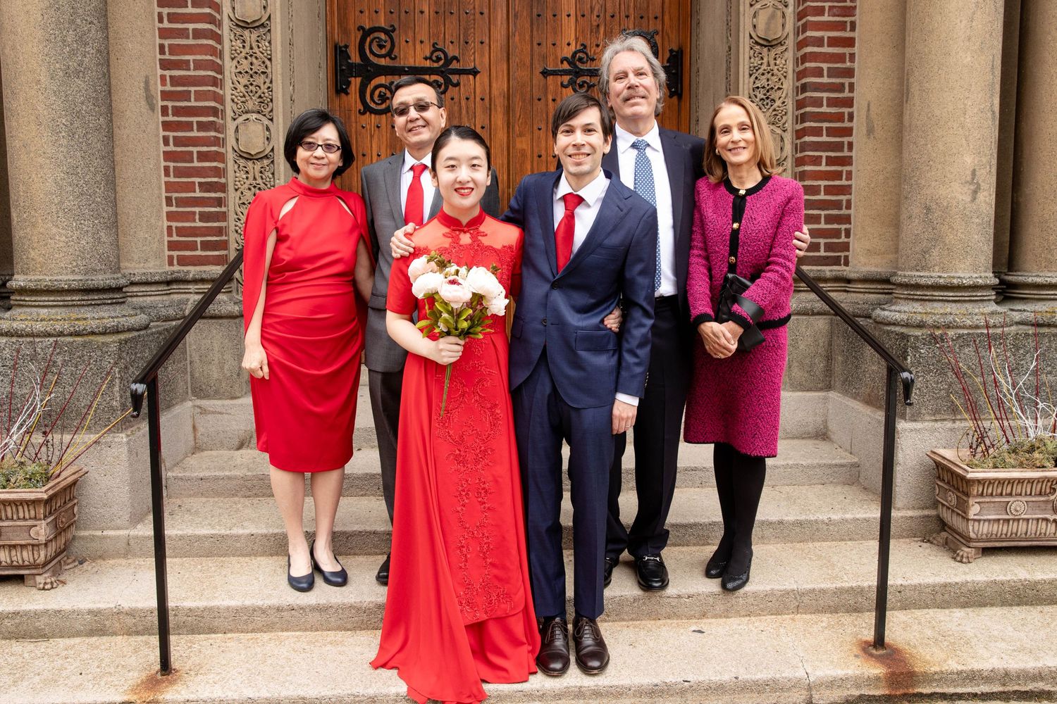 A wedding party poses on church steps with the couple in red and navy attire.